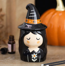 Load image into Gallery viewer, A glossy black witch figurine with a cartoon face, wearing a black hat with an orange band, displayed on a wooden surface with a blurred orange pumpkin and a small bottle in the background.