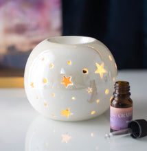 Load image into Gallery viewer, A white ceramic star-shaped wax melt burner diffuser or candle holder with glowing star cutouts, shown on a reflective white surface next to a small amber oil bottle with a purple label and dropper.
